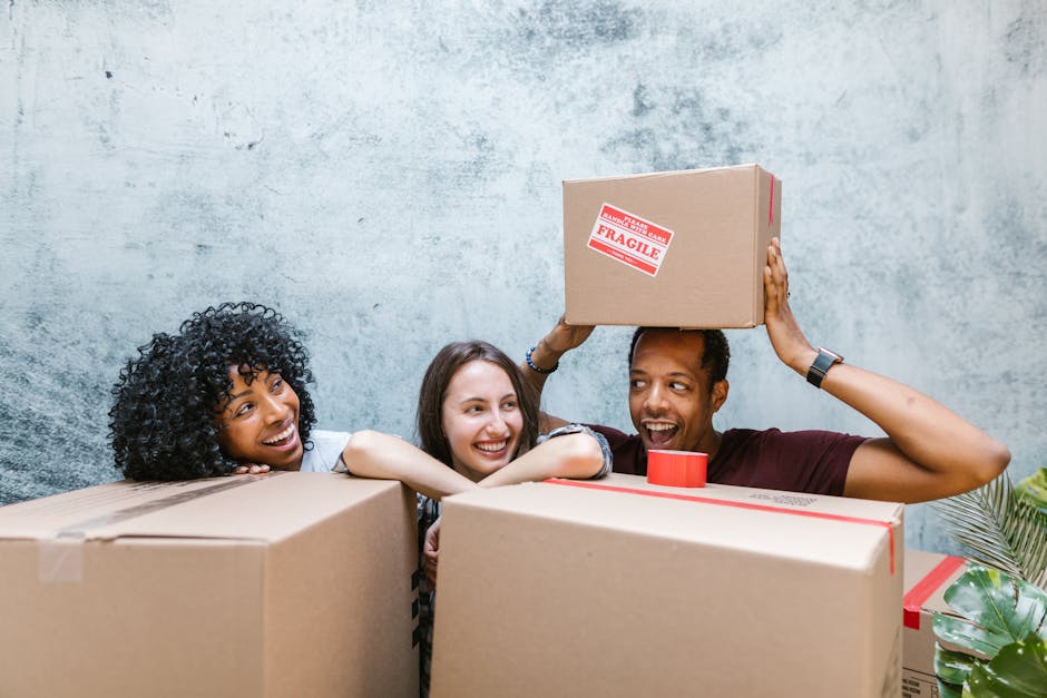 Three individuals, two women and one man, are engaged in a home relocation process indoors, surrounded by several large cardboard boxes. The woman on the left, with curly black hair, rests her chin on one of the boxes, smiling towards the camera. The woman in the middle, with straight dark hair, is leaning forward, also smiling, and appears to be observing the moving activity. The man on the right, with short dark hair, is holding a medium-sized cardboard box labeled 'Fragile' above his head, preparing to place it onto a surface or into a vehicle. The boxes are sealed with packing tape, with one box partially visible in front of the individuals. The background features a plain, textured wall with natural lighting, and a glimpse of a plant is visible in the lower right corner. This scene depicts the packing and handling aspects of furniture transport and home relocation services provided by Man with Van Becontree, emphasizing careful handling of fragile items during the moving process at a residential property.