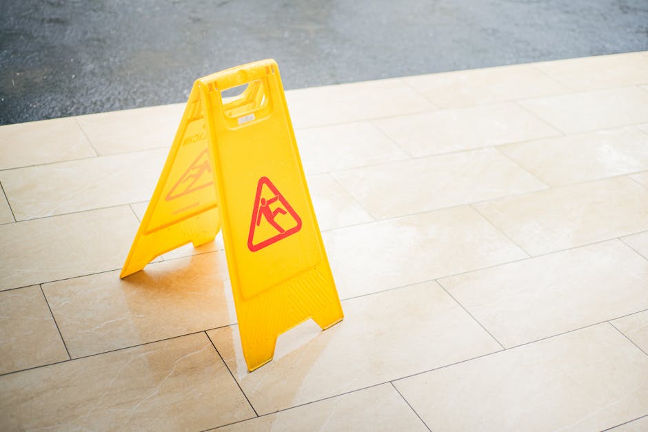 A yellow plastic wet floor warning sign standing on light beige tiled flooring inside or outside a property during a furniture transport or home relocation process. The sign displays a red and black icon of a person slipping, indicating a slippery surface, and is positioned near an area where packing and moving activities are underway. The background shows a smooth, paved surface outside the building, while the foreground features large, rectangular tiles with subtle textures. This image reflects the safety measures taken by Man with Van Becontree during removals and furniture transport tasks, emphasizing caution in moving logistics while handling boxes, furniture, or appliances inside or around a property.