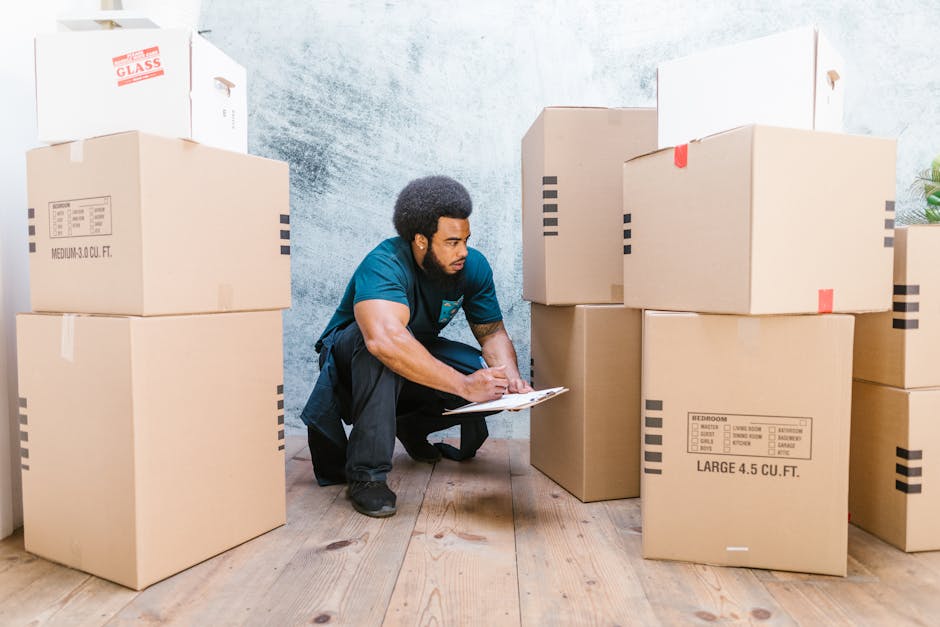 Three individuals, two women and one man, are engaged in a home relocation process indoors, surrounded by several large cardboard boxes. The woman on the left, with curly black hair, rests her chin on one of the boxes, smiling towards the camera. The woman in the middle, with straight dark hair, is leaning forward, also smiling, and appears to be observing the moving activity. The man on the right, with short dark hair, is holding a medium-sized cardboard box labeled 'Fragile' above his head, preparing to place it onto a surface or into a vehicle. The boxes are sealed with packing tape, with one box partially visible in front of the individuals. The background features a plain, textured wall with natural lighting, and a glimpse of a plant is visible in the lower right corner. This scene depicts the packing and handling aspects of furniture transport and home relocation services provided by Man with Van Becontree, emphasizing careful handling of fragile items during the moving process at a residential property.