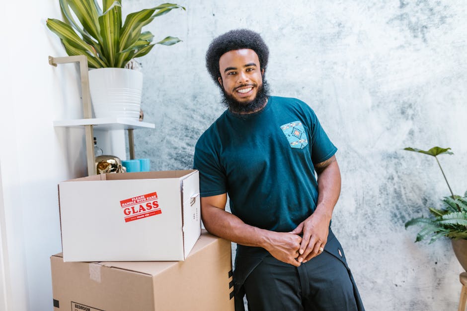 A man with dark skin, curly black hair, and a full beard is standing indoors next to stacks of cardboard moving boxes, one of which has a red 'GLASS' fragile sticker. He is smiling and wearing a blue t-shirt with a multicolored patch on the chest. The background features a light grey wall, a white shelf holding a large green houseplant in a white pot, and a small decorative object. To his right, there is a large potted plant with broad green leaves. The scene appears to be part of a home relocation or packing process, with the boxes arranged on a neutral flooring surface near a doorway or open space, and lighting is bright and even. The image reflects furniture transport and packing activities related to house removals, as handled by Man with Van Becontree, supporting efficient and organized home moving services.