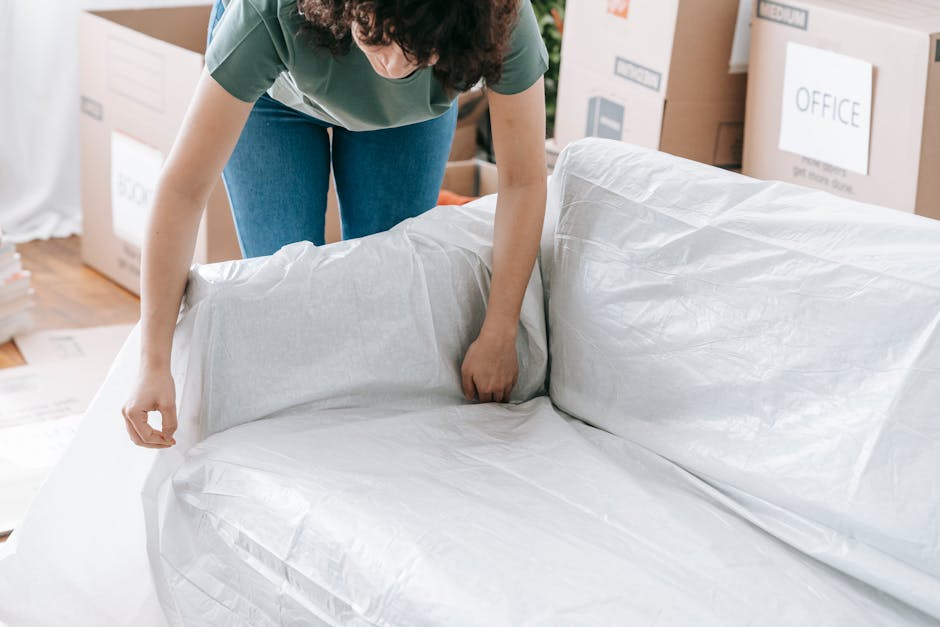 A person wearing a green t-shirt and blue jeans is seen during a home relocation process, wrapping a large piece of furniture with white protective plastic covering. The individual is inside a room filled with cardboard boxes, some labeled with items such as 'office'. The furniture appears to be a sofa or armchair, positioned on the floor and being carefully covered for transport. Nearby, moving equipment including a trolley and packing materials like cardboard boxes and bubble wrap are visible, indicating an organized packing and moving operation by a removal service. The setting is well-lit, with natural light illuminating the room, emphasizing the careful handling involved in furniture transport and packing during a professional house removal or relocation.