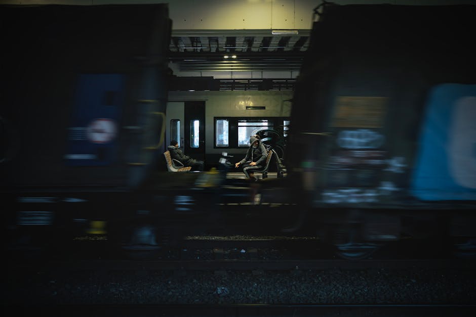 Two passengers seated inside a train carriage, visible through large windows, are waiting during a home relocation process. The interior features seating with light-colored upholstery, and the passengers are wearing face masks. Outside the carriage, briefly visible compartments and the platform's edge suggest the train is stationary at a station, possibly Becontree Station. The image captures a moment during general moving logistics, indicating a transport segment within a larger packing and moving operation. The scene is lit with natural daylight filtering through the windows, emphasizing the calm, routine environment of train travel amidst a house removal journey, as performed by Man with Van Becontree, a professional removals service specializing in furniture transport and home relocations.