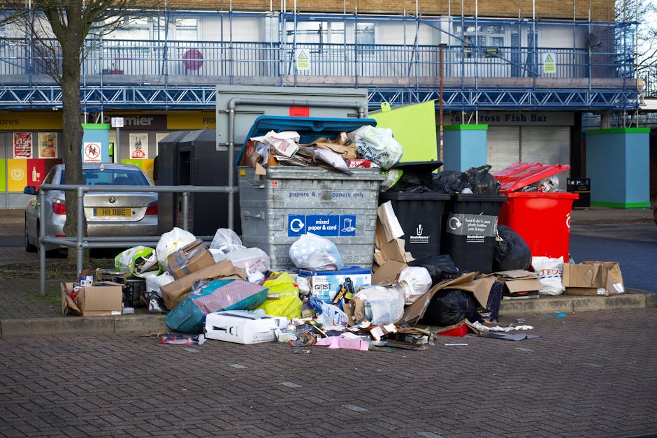 An outdoor scene showing multiple overflowing waste bins and rubbish bags placed on a paved sidewalk in front of a commercial building with closed shutters. The waste containers include a large grey mixed paper and cardboard bin with its lid open, filled with various packaging and paper waste; a black plastic bin with black rubbish bags piled beside it; and a red recycling bin. Loose cardboard boxes, paper, plastic, and other rubbish are scattered on the ground, some spilling out of the bins. To the left, a silver car is parked next to a metal railing, and behind the scene, a building with a blue framework, construction barriers, and a 'Cross Fish Bar' sign are visible. The scene is illuminated by natural daylight, and the setting highlights typical rubbish disposal outside a commercial or residential property, relevant to home relocation and moving services carried out by [COMPANY_NAME].