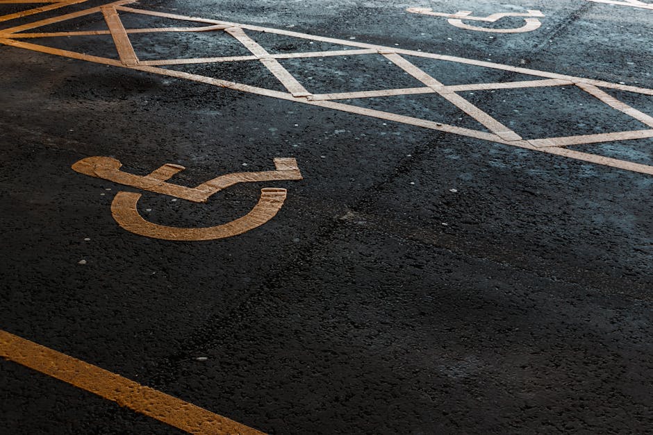 Close-up view of an outdoor parking area showing a yellow-painted accessible parking symbol on dark asphalt, with additional parking bay lines visible in the background. The surface appears slightly w