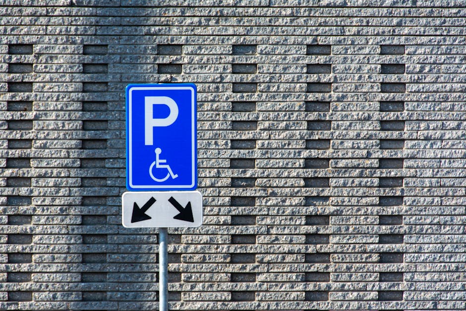 Close-up view of an outdoor parking area showing a yellow-painted accessible parking symbol on dark asphalt, with additional parking bay lines visible in the background. The surface appears slightly w