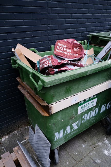 Two pedestrian trash bins positioned side by side on a sidewalk in an urban area. The left bin is dark green with a light blue lid, designated for recyclable waste, while the right bin is brown with a