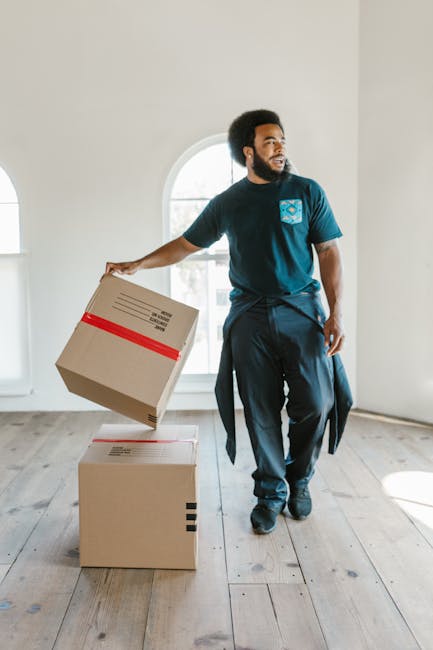 A man with dark curly hair and a beard is inside a bright, minimally furnished room with light-colored walls and a large arched window allowing natural daylight to illuminate the space. He is wearing a navy blue uniform with a light blue logo on the chest and is holding a cardboard box secured with red packing tape in his right hand, preparing to load or move it. In front of him are two additional cardboard boxes stacked on a wooden floor, used for home relocation and packing materials. Behind him, the interior features clean, smooth walls and a simple, modern ambiance, indicating an early stage of packing or furniture transport as part of a professional house removal service. The scene captures the process of furniture transport and packing during a moving task, with the man positioning the boxes. Man with Van Becontree, a company specializing in removals, is providing assistance in this context.