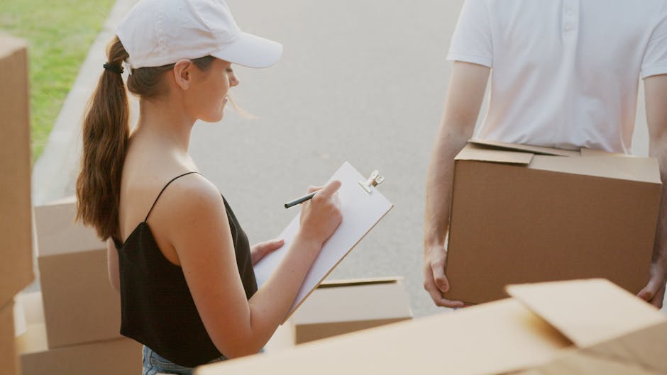 A young woman with light skin, long brown hair tied back in a ponytail, wearing a white cap, black sleeveless top, and jeans, is holding a clipboard and pen while standing outdoors on a pavement area near a residential property. She appears to be taking notes or recording information related to a home relocation or furniture transport process. In front of her, a partially visible man with light skin is wearing a white t-shirt and holding a medium-sized cardboard moving box, which is wrapped in brown packing tape. Several other cardboard boxes are stacked on the ground nearby, some sealed and others open or partially assembled. The scene is well-lit with natural light, and the background suggests a residential environment, aligning with the context of professional removals services offered by [COMPANY_NAME], such as packing and loading during a house move, as described on the webpage about certification and insurance for moving companies.
