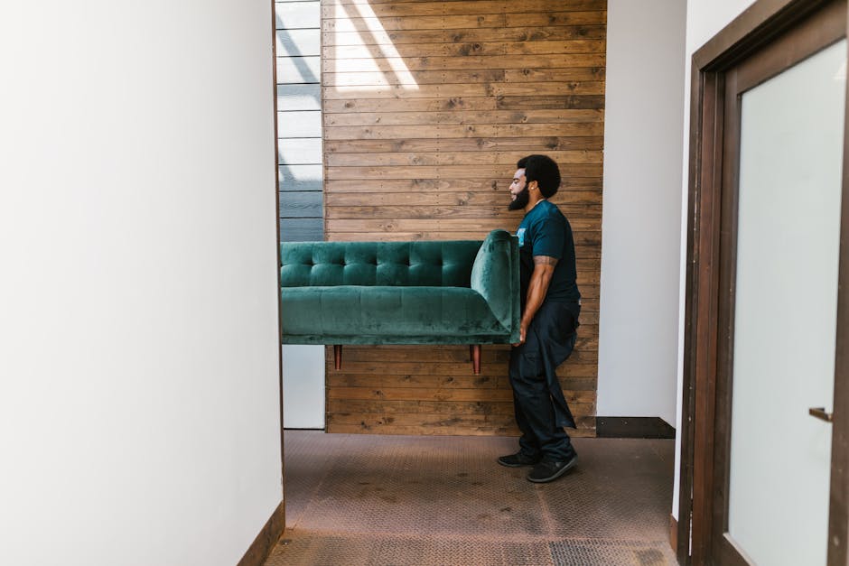 A man with dark curly hair and a beard is inside a bright, minimally furnished room with light-colored walls and a large arched window allowing natural daylight to illuminate the space. He is wearing a navy blue uniform with a light blue logo on the chest and is holding a cardboard box secured with red packing tape in his right hand, preparing to load or move it. In front of him are two additional cardboard boxes stacked on a wooden floor, used for home relocation and packing materials. Behind him, the interior features clean, smooth walls and a simple, modern ambiance, indicating an early stage of packing or furniture transport as part of a professional house removal service. The scene captures the process of furniture transport and packing during a moving task, with the man positioning the boxes. Man with Van Becontree, a company specializing in removals, is providing assistance in this context.