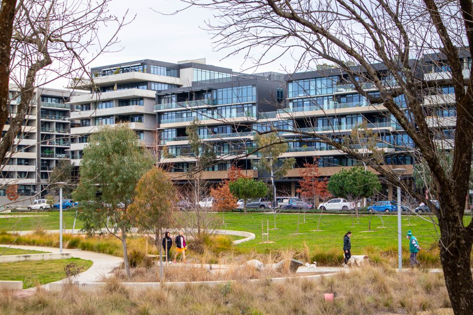 A scene of a residential building behind a public park during autumn, with multiple trees displaying green, yellow, and orange foliage. The park features a grassy area scattered with fallen leaves, an