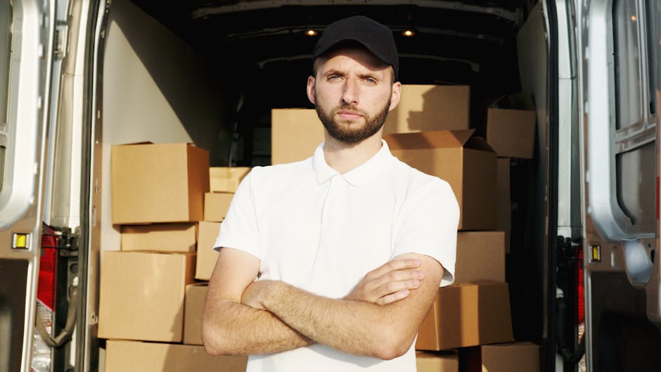 A young man with a serious expression, wearing a black cap and a white polo shirt, stands inside an open moving van loaded with numerous cardboard boxes of various sizes. The boxes, some sealed with packing tape and others partially wrapped in plastic or foam padding, are stacked neatly on the van's interior floor and sides, indicating a packing and loading process related to house relocation. The van's door is wide open, revealing a driveway or street outside, and the background shows additional parts of the vehicle's interior and external environment. The scene depicts a professional furniture transport and packing service, suggesting that [COMPANY_NAME] specializes in timely and organized home removal services such as same-day removals in Becontree, with an emphasis on logistics, safety, and efficient loading procedures.