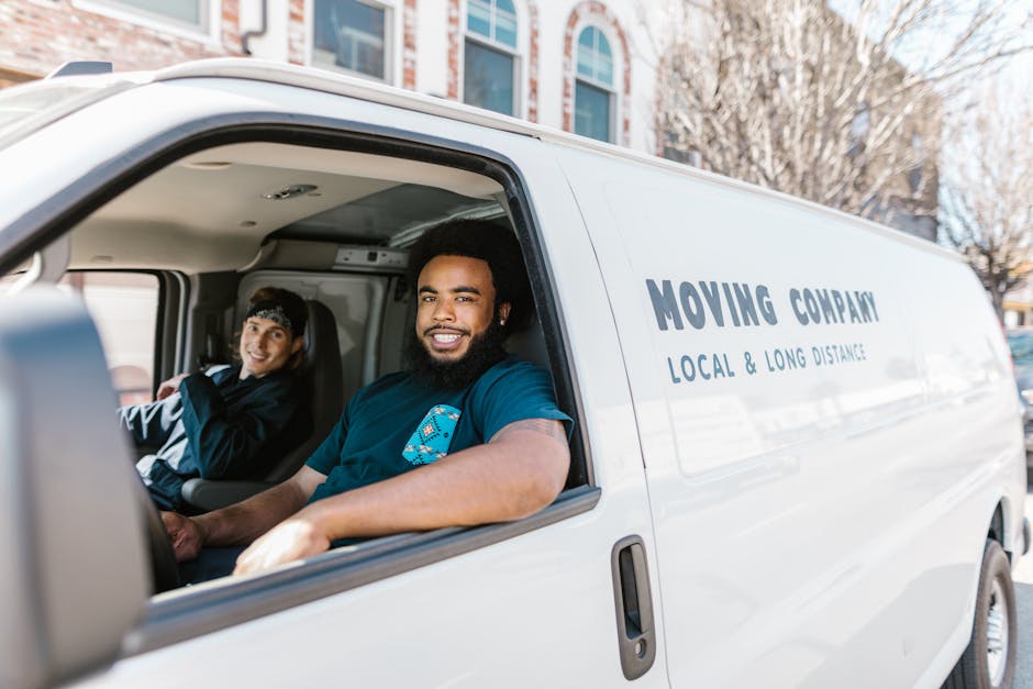 Inside a white moving van labeled 'Moving Company Local & Long Distance,' two male movers are visible, seated in the front cabin. The driver, with a full beard and dark curly hair, is smiling at the camera while wearing a navy blue t-shirt. The passenger, a woman with dark hair tucked under a black cap, is also smiling and looking slightly to the side. The van is parked outdoors on a street with residential buildings and leafless trees in the background, indicating a clear day. The interior of the van shows some packaging materials, including cardboard boxes and protective blankets, on the floor nearby, suggesting they are in the process of loading or unloading furniture and household items as part of home relocation or furniture transport services. The scene is well-lit, emphasizing the professional and organized nature of the moving process carried out by [COMPANY_NAME].