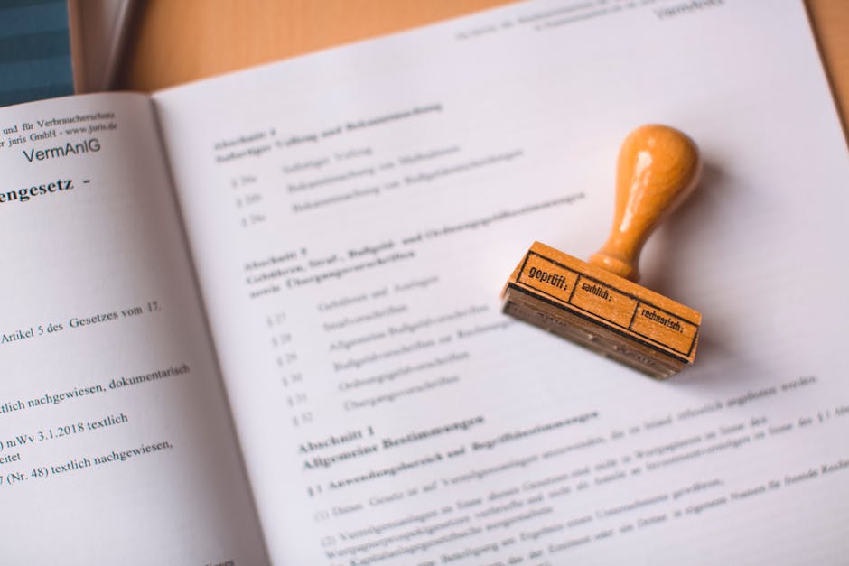 A close-up image shows an official document or legal paper laid open on a flat surface, with the focus on the left page which contains printed text in German. Resting on the right page is a rubber stamp with a wooden handle and a rubber base, featuring markings that include the words 'geprüft' (tested), 'schnitt' (cut), and 'reparatur' (repair) in black ink. The document appears to be related to legal or formal matters, with the paper slightly curled at the edges. The background is blurred and neutral, emphasizing the stamp and the document, which are associated with the process of official verification or approval. In the context of house removals and relocation services by Man with Van Becontree, this image visually represents the importance of proper documentation and permits involved in property relocation or compliance processes.
